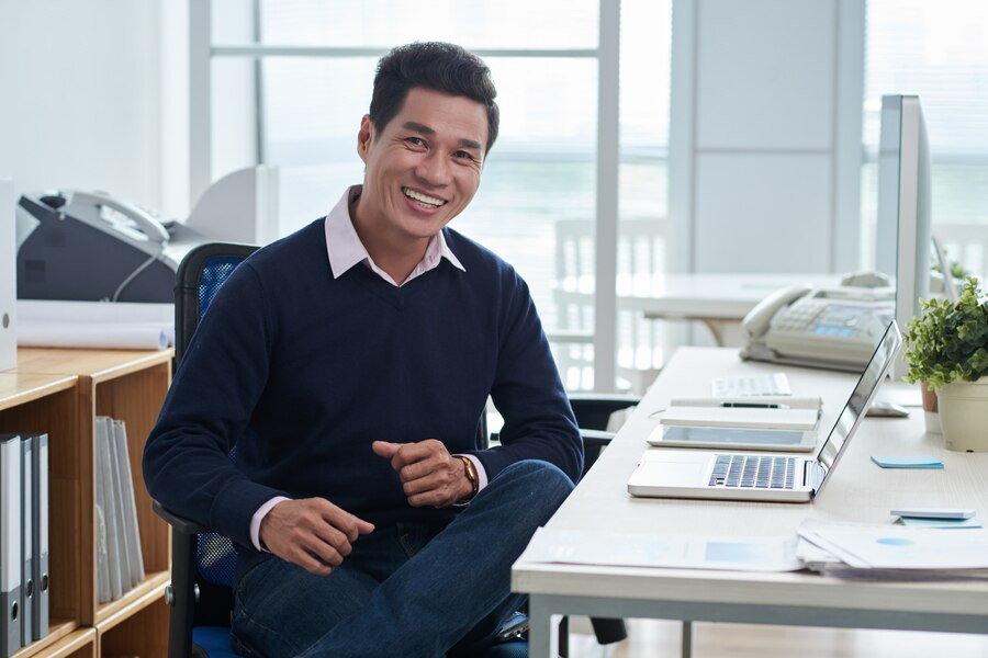 smiling-asian-man-sitting-desk-front-laptop-office-looking-camera-1098-20487