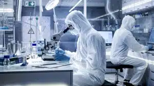 In A Secure High Level Laboratory Scientists In A Coverall Conducting A Research. Biologist Adjusts Samples In A Petri Dish With Pincers And Examines Them Under Microscope And His Colleague Analyzes Results On A Computer.